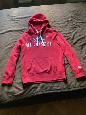 Röd Hollister hoodie med logga - Priset kan sänkas vid snabb affär. Snygg röd hoodie från Hollister med stor logga och fågeltryck på bröstet. Klassisk känguruficka framtill, vita dragsnören i huvan och en liten amerikansk flagga på ärmen. Perfekt för en chill och avslappnad stil.