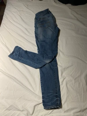 Blå slim fit jeans med slitningar - Säljer ett par blå slim fit jeans med snygga slitningar och klassisk femficksdesign. Jeansen har normal midja och raka ben, perfekta till en avslappnad stil. Materialet är mjuk bomull med lite stretch för extra komfort.