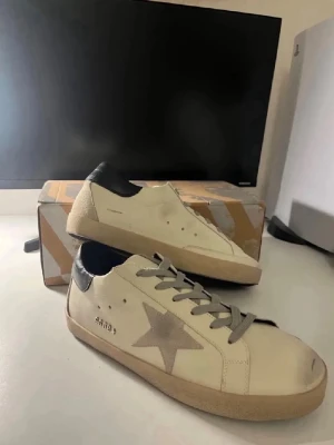 Vita sneakers med grå stjärna - Snygga vita sneakers med grå stjärndetalj på sidan och grå skosnören. Skorna har svart hälparti och beige sula. Klassisk låg modell med trendig design som passar till många olika stilar. Tillverkade i läder med detaljer i mocka.