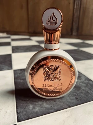 Lattafa Fakhar Eau de Parfum 100ml - Snygg parfymflaska från Lattafa i vitt och roséguld med rund form och dekorativt emblem framtill. Flaskan har en lyxig känsla med detaljer i metall och elegant kork. Perfekt för dig som gillar exklusiva dofter och stilren design. Det fins 2/3 kvar 