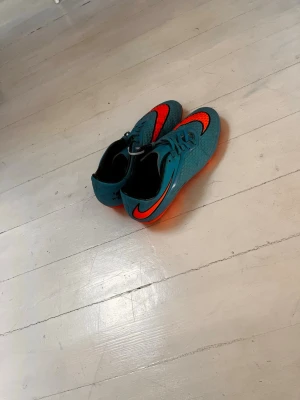 Blåa och orange Nike fotbollsskor - Säljer ett par snygga fotbollsskor från Nike i blått med orange detaljer och sula. Skorna har klassisk snörning och är tillverkade i syntetmaterial för bra grepp och kontroll på planen. Perfekta för dig som vill sticka ut på matchen!
