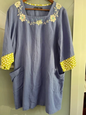 Blå linneklänning Gudrun Sjödén - Unik blå klänning från Gudrun Sjödén i linne med broderade blommor vid halsen och stora fickor. Ärmarna har gula muddar med vita prickar och trekvartsärm. Perfekt för dig som gillar färg och detaljer!