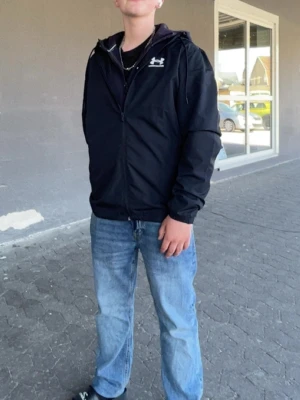 Svart vindjacka från Under Armour - Snygg svart vindjacka från Under Armour med dragkedja framtill och huva. Jackan har en diskret logga på bröstet och är tillverkad i ett lätt material som passar perfekt för blåsiga dagar. Enkel och stilren design som funkar till det mesta.