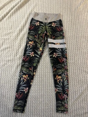 Mönstrade leggings med blommor från stronger - Snygga leggings med tropiskt blommönster i grönt, rosa och beige. Hög midja i ljusgrått och två vita ränder på ena benet. Stretchigt material som sitter tight och bekvämt. Perfekta för dig som vill sticka ut med färg och mönster.