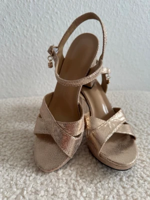 Nya. Guldiga sandaletter med klack - Säljer ett par snygga sandaletter i metallic guld med korsade remmar och öppen tå. Skorna har justerbar ankelrem med liten dekorativ detalj och en stadig klack. Materialet har en lätt skimrande struktur som ger extra lyxig känsla. Storlek 35 men stort i storleken och kan även passa 36cm, innelängd 23cm