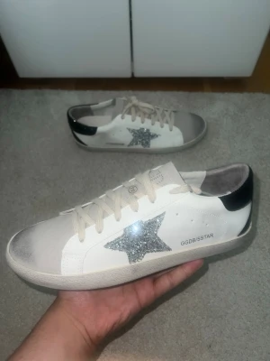 golden goose  - Säljer ett par vita sneakers med en silverglittrig stjärna på sidan och svart detalj på hälen. Skorna har vita snören och en klassisk låg profil. Märkningen GGDB/SSTAR syns på sidan. Perfekta för dig som vill sticka ut lite extra. Passar 44 o 43