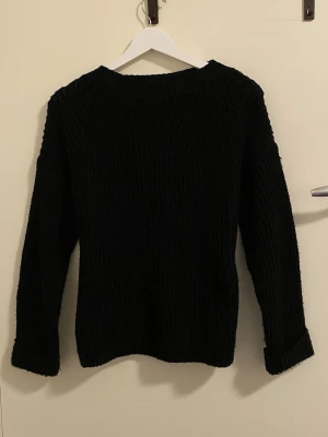 Svart stickad tröja med rund hals - En svart stickad tröja med rund hals och långa ärmar. Tröjan har en grovstickad struktur och uppvikta ärmslut. Perfekt för kyliga dagar och enkel att matcha med olika outfits.