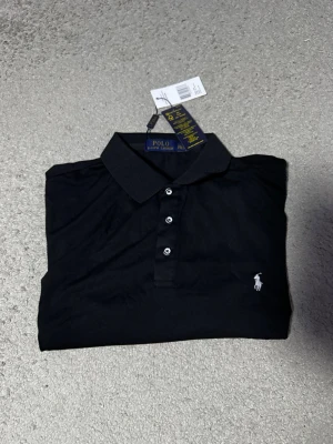 Svart pikétröja från Polo Ralph Lauren - Klassisk svart pikétröja från Polo Ralph Lauren med vit broderad logga på bröstet. Tröjan har korta ärmar, krage och knappar framtill. Tillverkad i mjuk bomull som känns skön mot huden. Perfekt för en clean och stilren look.