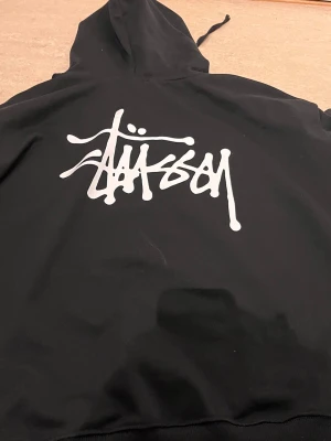 Svart hoodie från Stüssy med logga - Svart hoodie från Stüssy med stor vit logga på ryggen och mindre logga på bröstet. Klassisk känguruficka framtill, ribbade muddar och justerbar huva med dragsko. Perfekt för en streetwear-look och riktigt skön att bära.