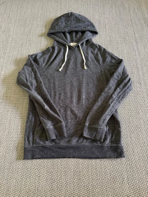 Mörkgrå hoodie från H&M L.O.G.G. - Mörkgrå hoodie från H&M:s L.O.G.G.-kollektion med vita dragsnören i huvan. Tillverkad i mjukt och tunt material, perfekt för en avslappnad stil. Långa ärmar och klassisk passform. Enkel och stilren design utan tryck eller mönster.