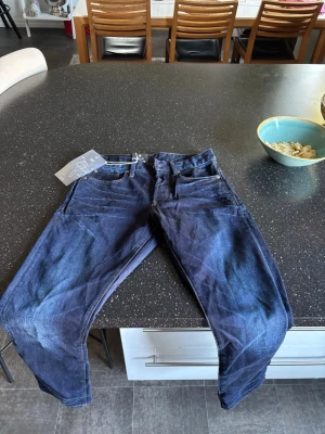 Mörkblå raka jeans från raw med tvättade detaljer - Säljer ett par mörkblå jeans med raka ben och snygga tvättade detaljer. Jeansen har klassisk femficksmodell och är tillverkade i denim. Perfekta för dig som gillar en stilren och avslappnad look. I storleken 29/32