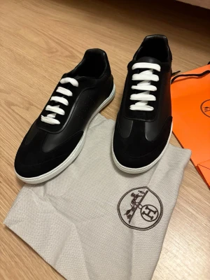 Svarta Hermes sneakers med vita sulor - Snygga svarta sneakers från Hermès med vita sulor och vita skosnören. Skorna har en stilren design i läder och mocka, samt diskreta perforerade detaljer på sidan. Perfekta för dig som vill ha en exklusiv och modern look.