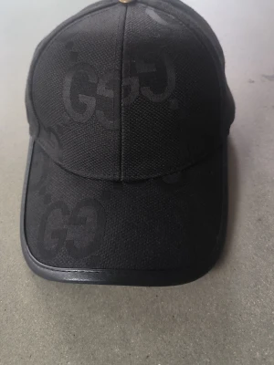 Svart keps med GG-mönster - Snygg svart keps med ton-i-ton GG-mönster och böjd skärm. Kepsen har en liten metallknapp på toppen och är tillverkad i ett tåligt tygmaterial. Perfekt accessoar för en trendig och sportig look.