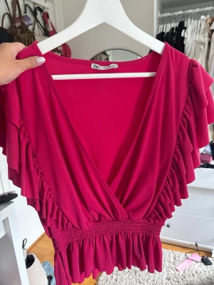 Rosa omlottopp med volanger från ZARA - Supersnygg rosa omlottopp från ZARA med bred resår i midjan och volangdetaljer längs ärmarna. Djup v-ringning framtill och luftig passform som ger en trendig look. Perfekt att styla med jeans eller kjol för en cool vibe.