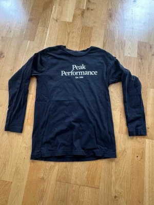Svart långärmad Peak Performance t-shirt - 150cl. Svart långärmad t-shirt från Peak Performance med vit logga och text på bröstet. Klassisk rund halsringning och rak passform. Perfekt för en avslappnad och stilren look.Nyskick, bara provad.