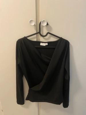 Svart omlottopp med lång ärm - Stilren svart omlottopp med lång ärm och djup v-ringning. Toppen har en smickrande omlottdesign framtill och är tillverkad i ett mjukt, stretchigt material. Perfekt att styla med jeans eller kjol för en trendig look. 