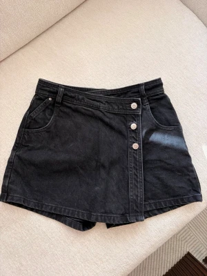 Svarta jeans shorts med knappar - Snygg svart shorts i denim med omlott-look framtill och tre silverfärgade knappar. Klassisk femficksmodell med bälteshällor och raka ben. Perfekt för dig som vill ha kjol-looken men ändå känna dig bekväm i shorts. Första ägare. Silverdetaljer har ändrat i färg, se bilder.