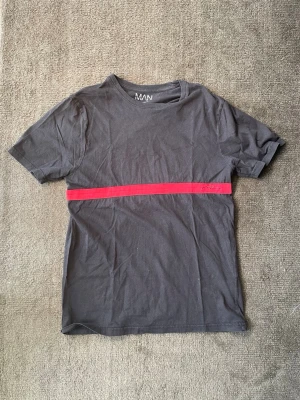 T-shirt - BoohooMan - Snygg svart t-shirt från MAN med  röd rand över bröstet.  ✅ Svart bomullstyg, avslappnad passform ✅ Röd rand tvärs över bröstet med broderad “Man”-logga ✅ Rund halsringning ✅ Korta ärmar  Skick: Mycket bra Storlek: L  Enkel och snygg basic-t-shirt med en cool detalj – perfekt att kombinera med det mesta i garderoben. Skriv gärna vid frågor!