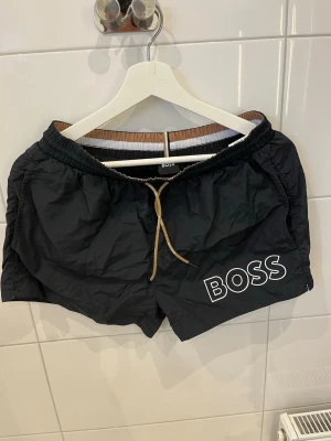 Svarta badbyxor från BOSS - Snygga svarta badbyxor från BOSS med vit logga på vänster ben. De har elastisk midja med beige dragsko och sidofickor. Perfekta för stranden eller poolen och har en klassisk, sportig look.