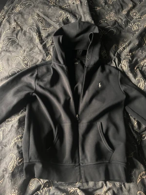 Svart hoodie från Polo Ralph Lauren - Snygg svart hoodie från Polo Ralph Lauren med dragkedja framtill. Klassisk huva och den ikoniska loggan broderad på bröstet. Perfekt för en chill och stilren look.