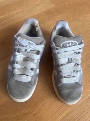 Gråa Adidas sneakers i mocka - Gråa Adidas sneakers med vita detaljer och breda vita skosnören. Skorna har klassiska tre ränder på sidan och Adidas-logga på plösen. Ovandelen är i mocka och sulan är vit. Perfekta för en avslappnad streetwear-look.
