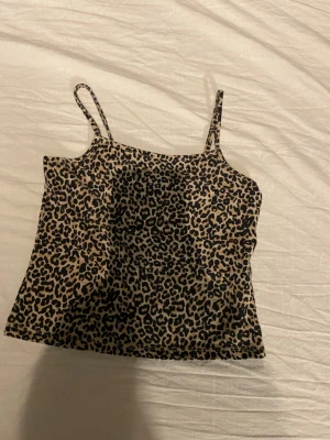 Leopardmönstrat linne med smala band - Säljer ett leopardmönstrat linne med smala axelband. Linnet är i beige och svart med ett klassiskt djurmönster och har en rak, kort passform. Perfekt för dig som vill sticka ut lite extra med din outfit.