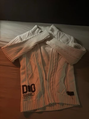 Unik Stickad Tröja - Snygg vit stickad tröja med dragkedja, klassiskt kabelstickat mönster och en broderad D&G-logga nertill. Perfekt inför hösten!