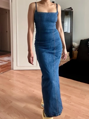 Blå lång jeansklänning från Bershka - Snygg lång jeansklänning från Bershka i blå denim. Klänningen har smala axelband, figurnära passform och markerade sömmar. Perfekt för dig som gillar en stilren och trendig look med denimkänsla.