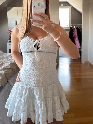 Vit spetsklänning från Gina Tricot - Supersöt vit kort klänning från Gina Tricot i spets med volangkant och knytning vid bysten. Klänningen har smala axelband och ett romantiskt mönster i tyget. Perfekt för sommarens alla tillfällen!