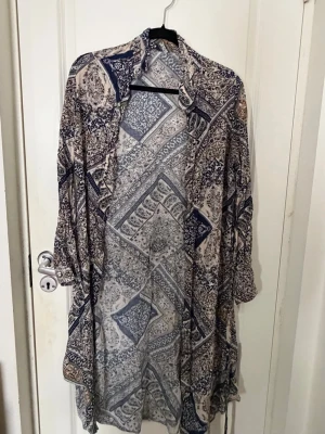 Mönstrad strandkimono i blått och beige - Luftig strandkimono med bohemiskt mönster i blått och beige. Lång och öppen modell med långa ärmar och dragsko i midjan. Perfekt att slänga över bikinin på stranden för en avslappnad och snygg look.