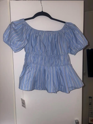 Blå- och vitrandig blus med puffärm - Supersöt blus med blå och vita ränder, puffiga korta ärmar och smock i midjan. Blusen har en luftig passform och en lätt volang nedtill. Perfekt för dig som gillar en feminin och trendig stil.