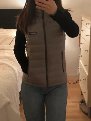 Äkta Grå Patagonia Dunväst  - Snygg, knappt använda, grå dunväst från Patagonia med quiltad design och hög krage. Västen har två dragkedjeförsedda fickor på sidorna och stängs med dragkedja framtill. Perfekt att slänga över en hoodie eller långärmad tröja för extra värme. Kan även passa XS