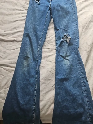 Blå bootcut jeans med slitningar  - Säljer ett par blå bootcut jeans med slitningar på låret och vid fickan. Jeansen har en klassisk femficksmodell och är tillverkade i jeansmaterial. Perfekta för dig som gillar en retro och avslappnad stil.