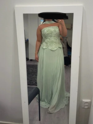 Ljusgrön långklänning  - Elegant ljusgrön korsett långklänning med plisserad kjol. Klänningen är använd enbart ett par timmar. Perfekt för bal eller speciella tillfällen. Med klänningen medföljer dess skarf runt halsen som är avtagbar