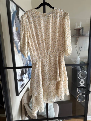 Blommig kortklänning från Adoore - Supersöt kortklänning från Adoore i vitt med små gula blommor och gröna detaljer. Klänningen har asymmetrisk volangkant, rund halsringning och trekvartsärm. Perfekt för sommarens alla tillfällen!