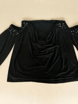 Svart offshoulder topp med pärlor - Stilren svart offshoulder topp med långa ärmar och pärldetaljer vid axlarna. Toppen har en snygg draperad front och är tillverkad i ett glansigt, mjukt material som ger en elegant look.
