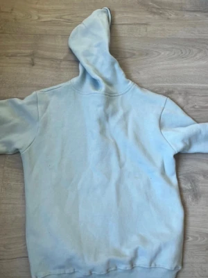 Ljusblå hoodie med strass och tryck - Säljer en ljusblå hoodie med dragkedja och huva. På framsidan finns ett coolt tryck med texten 'AIM FOR THE MOON' i strass och en tecknad figur med vingar och gevär, samt en jordglob. Hoodien har ribbade muddar och är i mjukt material.