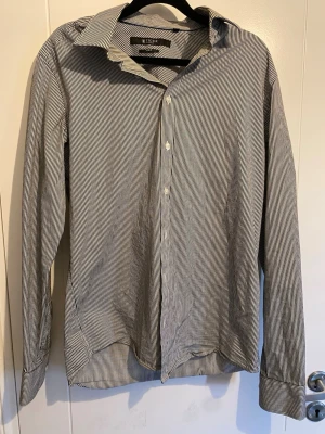 Randig skjorta från Tiger of Sweden - Snygg vit och svart randig skjorta från Tiger of Sweden i slim fit-modell. Skjortan har klassisk krage, knäppning framtill och långa ärmar. Perfekt för dig som gillar stilrena och tidlösa plagg.