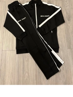 Palm Angels svarta mjukisbyxor - Svarta mjukisbyxor från Palm Angels med vita sidoränder och logga på benet. Byxorna har resår i midjan och dragkedja vid benslut. Tillverkade i ett mjukt och stretchigt material som ger en sportig och trendig look.