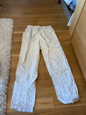 Beiga linnebyxor med dragsko - Säljer ett par ljusbeige linnebyxor med avslappnad passform och resår i midjan samt dragsko för justerbarhet. Byxorna har raka ben och är tillverkade i ett luftigt linnematerial, perfekta för varma dagar.