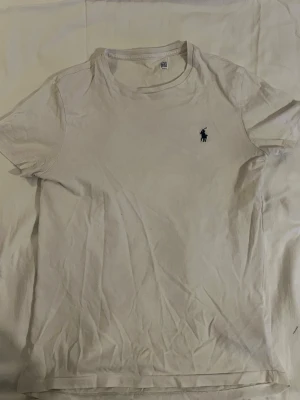 Vit t-shirt från Ralph Lauren - Klassisk vit t-shirt från Ralph Lauren med rund halsringning och den ikoniska broderade loggan i mörkblått på bröstet. Tillverkad i mjukt bomullsmaterial och har en enkel, stilren design som passar till det mesta.