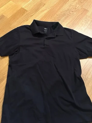 Svart pikétröja från Uniqlo - Klassisk svart pikétröja från Uniqlo med krage och korta ärmar. Tröjan har två knappar vid halsen och är tillverkad i ett mjukt och bekvämt material som passar perfekt till alla tillfällen.