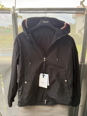   Moncler windbreaker - Snygg svart vindjacka från Moncler med huva och dragsko. Jackan har två framfickor med tryckknappar och dragkedja framtill. Insidan av huvan har en röd, vit och blå randig detalj. Perfekt för blåsiga dagar och stilren look. Perfekt till hösten!😍💯