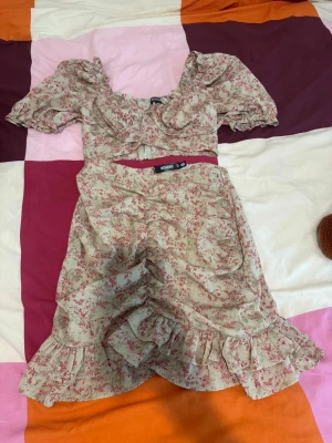 Blommig kortklänning med volang - Söt kortklänning med ljusbeige botten och rosa blommigt mönster. Klänningen har puffiga korta ärmar, v-ringad urringning och volangdetaljer vid nederkanten. Tillverkad i ett lätt bomullstyg, perfekt för varma dagar.