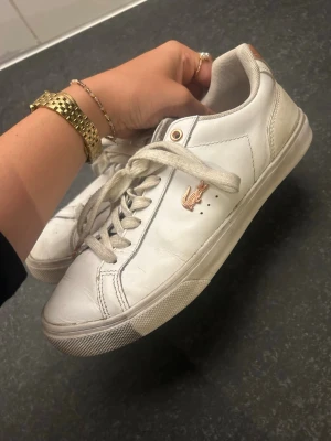 Vita Lacoste sneakers i läder - Säljer ett par klassiska vita sneakers från Lacoste i läder med gulddetaljer och den ikoniska krokodilloggan på sidan. Skorna har vita snören, diskreta sömmar och låg profil som passar till många olika stilar.