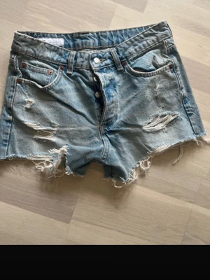 Blå denimshorts med slitningar - Säljer ett par blå jeansshorts i klassisk denim med råa, fransiga kanter och slitna detaljer framtill. Shortsen har fem fickor, bälteshällor och knäppning med knappar. Perfekta för en avslappnad och trendig look.
