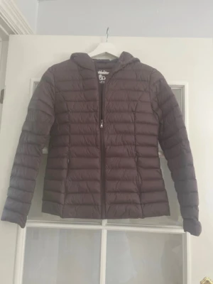 Mörklila dunjacka med huva från JOTT - Snygg mörklila dunjacka från JOTT med huva och dragkedja framtill. Jackan har ett quiltat mönster och en patch med JOTT-loggan på ärmen. Perfekt för kalla vinterdagar och riktigt lätt att bära.