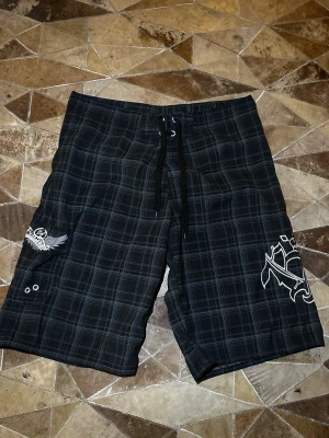 Svarta rutiga shorts med snörning - Säljer ett par svarta shorts med rutigt mönster och vita tryckdetaljer på benen. Shortsen har snörning i midjan och en avslappnad passform, perfekta för sommardagar. Materialet känns lätt och bekvämt, och designen har en streetwear-vibe.