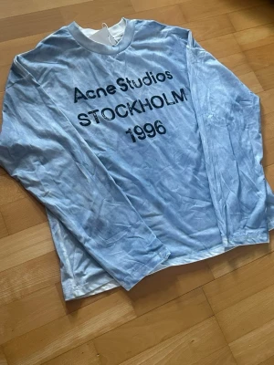 Long-sleeve T-shirt Acne Studios Stockholm 1996  - Denna äkta långärmade t-shirt från Acne Studios har en karaktäristisk ljusblå tvättad finish som ger den en vintage-känsla. På framsidan syns ett tydligt tryck i svart med texten ”Acne Studios STOCKHOLM 1996”, vilket hyllar varumärkets arv.   Skick: 9/10  Färg: Ljusblå (tvättad)  Snabb och omsorgsfull frakt