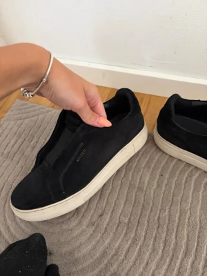 Svarta sneakers från Axel Arigato - Stilrena svarta sneakers från Axel Arigato i mocka med vit sula. Diskret logga i guld på sidan och minimalistisk design utan snörning. Passar perfekt till en clean och modern look.